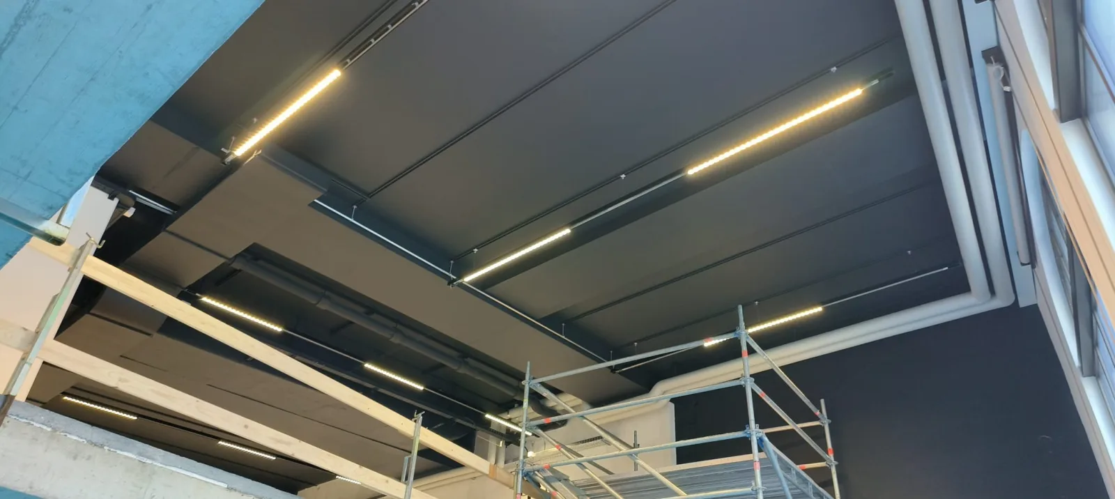 Black acoustic ceiling panels and warm LED strip lights mounted overhead, with scaffolding below