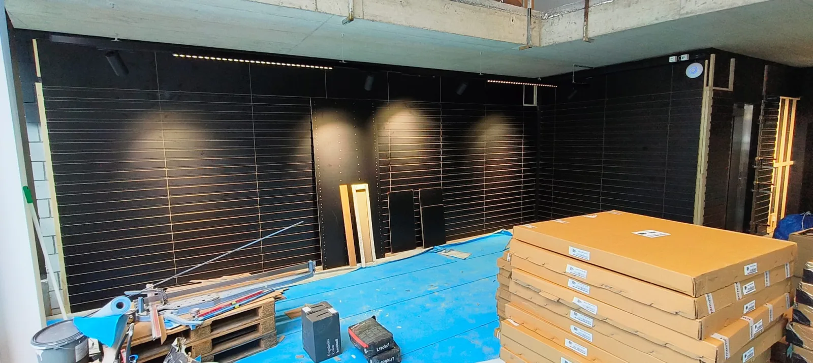 Black slatwall wall with stacks of flat-packed shelf panels and building materials on a pallet in front