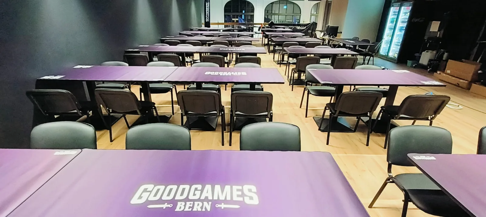 The tournament hall fully set up with rows of tables in purple GoodGames Bern playmats, chairs and a stocked drinks fridge
